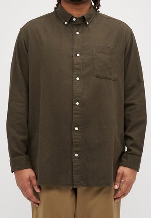 Uomo che indossa una camicia a maniche lunghe color oliva scuro con taschino sul petto e colletto abbottonato, abbinata a pantaloni marrone chiaro su sfondo neutro.