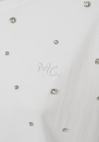 White shirt featuring a subtle “MG” embroidered logo, adorned with scattered silver rhinestones across the fabric. Soft texture, casual design.