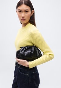 Μαύρο ψεύτικο δερμάτινο clutch με σούρες, κρατημένο από μοντέλο που φορεί κίτρινο πουλόβερ με ραβδώσεις και σκούρο τζιν παντελόνι.