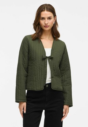 Vrouw in een olijfgroene gewatteerde jas die aan de voorkant is vastgebonden over een witte top en zwarte broek, die recht vooruit kijkt met een ontspannen uitdrukking.