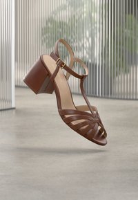 Sandales à talons en cuir marron avec des lanières croisées, un design à bout ouvert, une bride à la cheville et un talon épais, présentées sur un fond minimaliste.