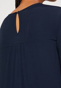 Gros plan sur le dos d'un chemisier bleu foncé avec une petite ouverture en forme de goutte d'eau fermée par un bouton noir près de l'encolure.