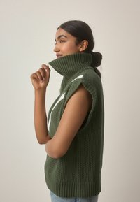 Grüner gestrickter Weste mit hohem Rippkragen und offenen Schultern; verfügt über ein weißes geometrisches Muster; weiche Textur; lockere Passform.