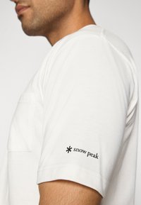 T-shirt bianco in cotone con scollatura a girocollo e taschino. Presenta il logo nero "snow peak" sulla manica sinistra, texture liscia e maniche corte.