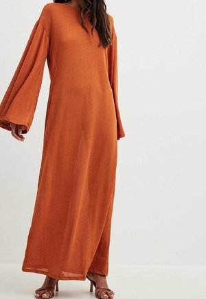 Woman wearing a long, loose-fitting burnt orange dress with wide sleeves and brown heeled sandals against a plain white background.