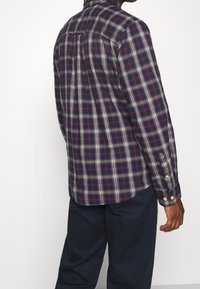 Camicia a maniche lunghe in plaid nei colori blu navy, rosso e crema, con colletto classico, orlo curvo e polsini con bottone, arricchiti da un delicato dettaglio a piega sul retro.