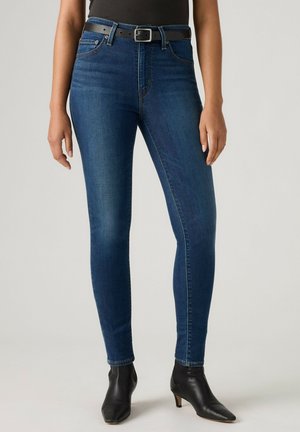 Frau, die blaue Skinny Jeans mit schwarzem Gürtel und schwarzen Stiefeletten trägt, steht vor einfachem hellem Hintergrund.