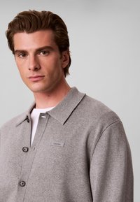 Jeune homme aux cheveux bruns coiffés, portant une veste-chemise gris clair boutonnée sur une chemise blanche, sur un fond clair uni.