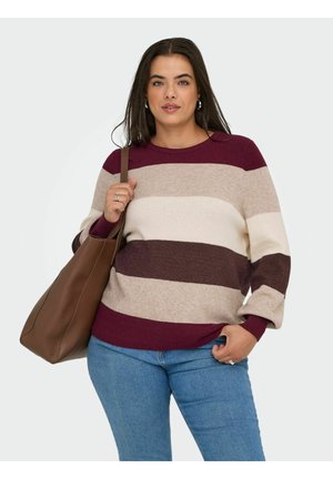 Frau trägt einen gestreiften Pullover in Bordeaux, Beige, Creme und Braun, blaue Jeans und hält eine große braune Tragetasche über der Schulter.