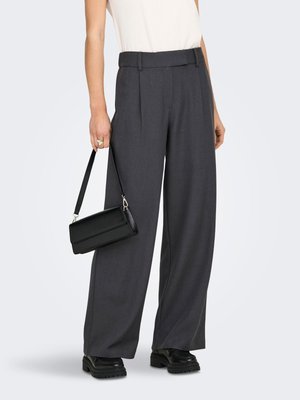 Osoba koja nosi široke hlače tamnosive boje, crne masivne cipele, bijelu majicu, držeći malu crnu torbicu za remen lijevom rukom.