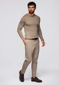 Vaaleanruskea villapaita pitkillä hihoilla, yhdistettynä vaaleanbeigeihin säädettyihin housuihin ja tummanruskeisiin kenkiin, jotka ovat muotoiltu tiukasti istuviksi.