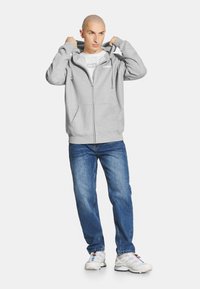 Szara bluza na zamek z dużymi kieszeniami z przodu, z troczkami na kapturze, noszona na białym t-shircie, w połączeniu z niebieskimi dżinsami i białymi trampkami.