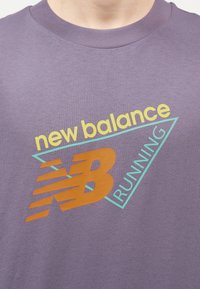 Μωβ πουκάμισο με λογότυπο της New Balance σε κίτρινο, πορτοκαλί και τιρκουάζ, που περιέχει τις λέξεις «new balance» και «RUNNING» μέσα σε ένα τριγωνικό σχήμα.