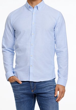 Man die een lichtblauw overhemd met lange mouwen en een spijkerbroek draagt, staand met één hand in de zak.