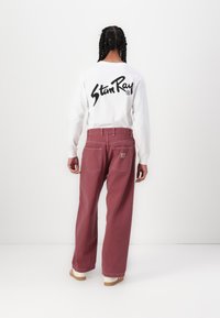 Pikendate valge T-särk pikkade varrukatega, mille seljal on must "Stan Ray" logo, ühendatud laia burgundia värvi pükste ja heledast puidust platvormsandaalidega.