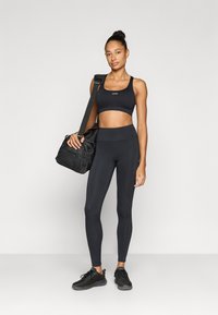 Μαύρο αθλητικό σουτιέν και κολάν με ψηλή μέση από ελαστικό ύφασμα. Το μοντέλο κρατάει μια μαύρη αθλητική τσάντα και φοράει μαύρα αθλητικά παπούτσια.