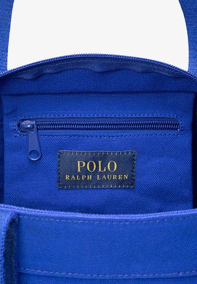 Εσωτερικό μπλε υφασμάτινης τσάντας με τσέπη με φερμουάρ και μια ετικέτα που γράφει "POLO RALPH LAUREN" με χρυσά γράμματα σε σκούρο φόντο.