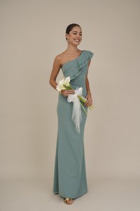 One-shoulder teal gown with ruffle, fitted silhouette, smooth texture. Holds a bouquet of white flowers tied with a sheer ribbon. Gold sandals.