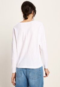 Frau mit dunkelhaarigen Haaren in einem lockeren Dutt trägt ein weißes Langarmoberteil und blue Denim-Jeans, steht mit dem Rücken zu einem einfarbigen Hintergrund.