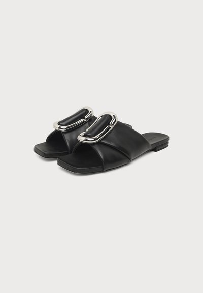 Sandales plates en cuir noir avec des brides larges et de grandes boucles ovales en métal argenté sur fond blanc.