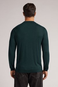 Ciemnozielony sweter dzianinowy z okrągłym dekoltem, długimi rękawami i luźnym krojem, zestawiony z szarymi dżinsami. Widoczny szczegół teksturowanej tkaniny.