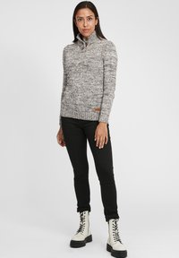 Tekstureret grå og hvid strikket sweater med høj krave og dekorative knapper, båret med sorte bukser og cremede snørebåndsstøvler.
