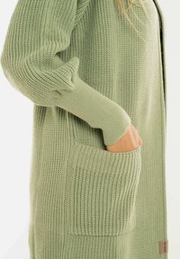 De hand van een persoon in de voorkant van een lichtgroene gebreide cardigan met textuurstof en lange mouwen.