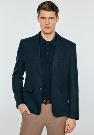 Młody mężczyzna ubrany w granatową marynarkę na ciemnogranatowej koszulce polo i beżowych spodniach z brązowym paskiem, stojący z jedną ręką w kieszeni.