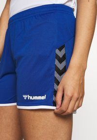 Blå sportshorts med vit fåll, med en svart sidopanel med chevronmönster och Hummel-logotyp i vitt på framsidan.