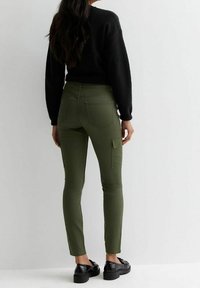 Pantalones cargo verde oliva de corte slim, con bolsillos frontales y traseros. Llevado con un suéter negro y zapatos de cordones negros.