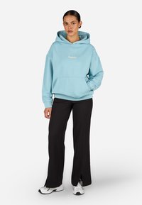 Ανοιχτό μπλε φούτερ με καγκουρό τσέπη και κουκούλα με κορδόνι, με το λογότυπο 'Pegador'. Συνδυάζεται με μαύρο παντελόνι φαρδύ και λευκά αθλητικά παπούτσια.