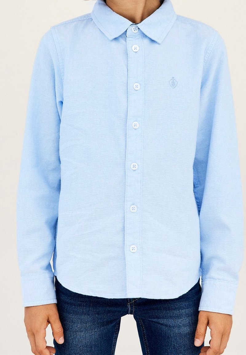 Enfant portant une chemise bleu clair à manches longues avec col et un jean bleu foncé, debout les bras détendus le long du corps.