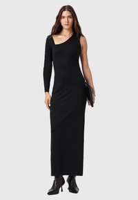 Musta, yhdellä olkapäällä varustettu mekko, jossa on vartalonmyötäinen siluetti ja miellyttävän kiiltävä, sileä kangas. Malli pitelee mustaa clutch-laukkua ja käyttää nilkkureita.