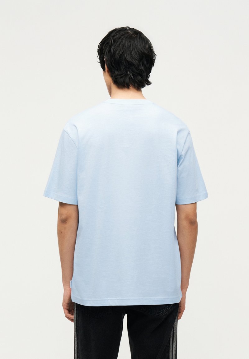 Hombre con cabello negro que lleva una camiseta azul claro lisa de manga corta y pantalones negros, de pie de espaldas sobre un fondo liso.