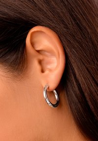 Boucle d'oreille en argent avec une finition polie, de forme circulaire, et un petit détail de charnière, portée sur le lobe. Les cheveux couvrent partiellement l'oreille.