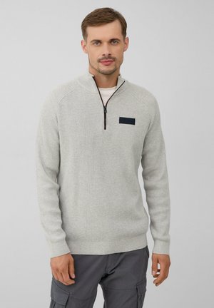 Lichtgrijze gebreide pullover met een ritskraag, voorzien van een textuurpatroon en een donkere vlek op de linkerkant van de borst. Gekleed met grijze cargobroek.