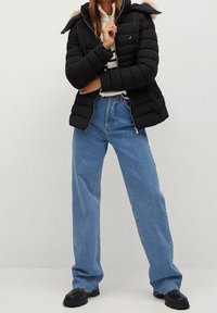 Zwarte pufferjack met een bontgevoerde capuchon, ritssluiting aan de voorkant, gecombineerd met lichtblauwe wijde spijkerbroeken en zwarte chunky schoenen. Soepele texturen en een casual pasvorm.