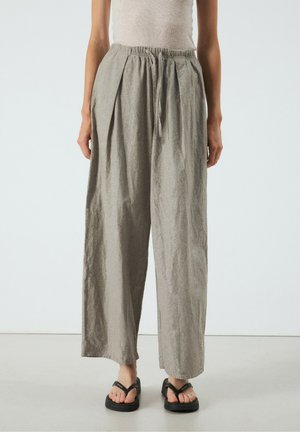 Femme portant un pantalon ample gris clair à cordon et des tongs plateformes noires, debout devant un fond uni et clair.