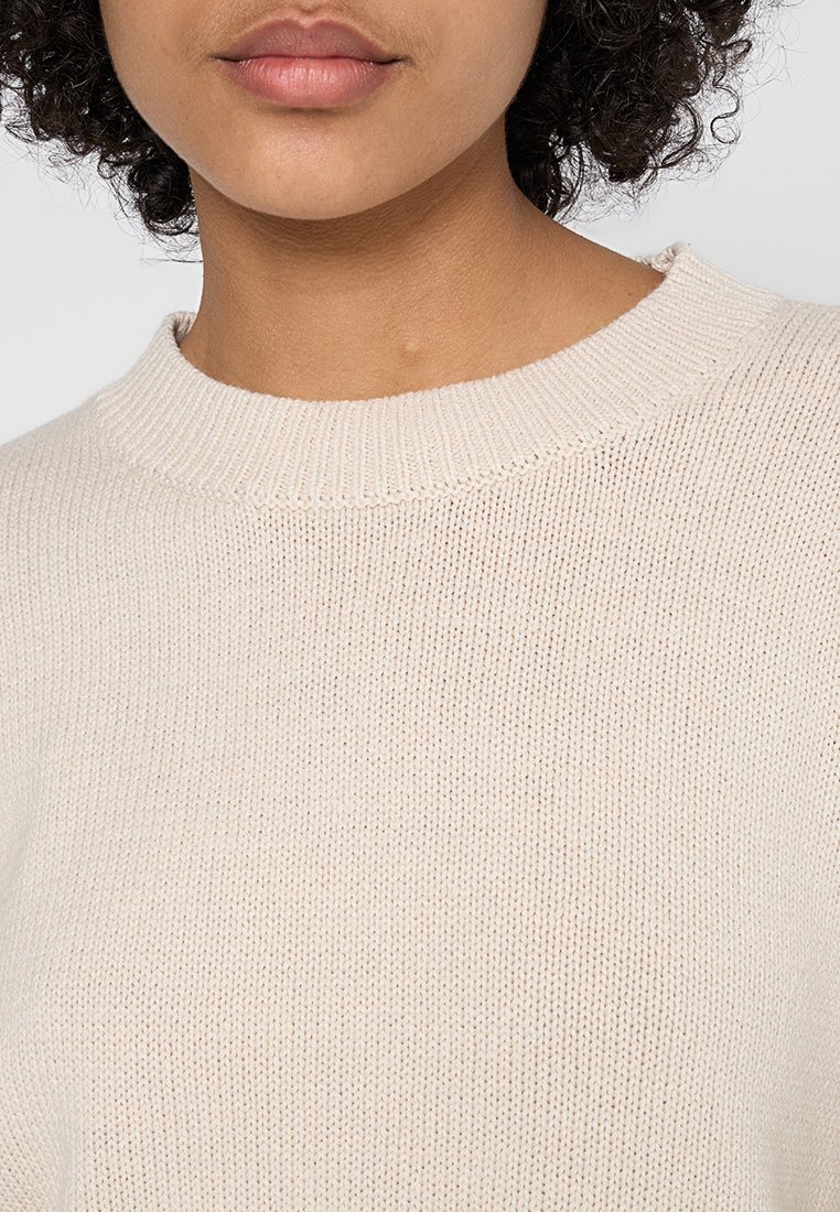 Maglione beige a maglia con scollatura rotonda, caratterizzato da un motivo in tessuto testurizzato e bordi a coste, visibile dalla zona della parte superiore del torace.