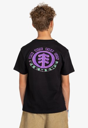 Młoda osoba z kręconymi włosami ubrana w czarną koszulkę z fioletowo-zielonym okrągłym logo "Element" oraz tekstem odnoszącym się do wiatru, wody, ognia i ziemi.