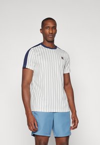 Mann iført en hvit-og-marine-stripete kortermet skjorte og lyseblå shorts, stående mot en ensfarget lys bakgrunn.