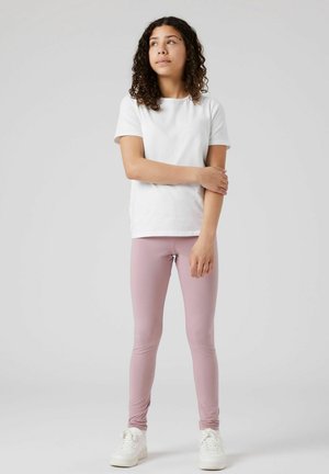 Jeune fille avec des cheveux bouclés se tient debout, portant un t-shirt blanc, des leggings mauves et des baskets blanches, regardant légèrement sur le côté.