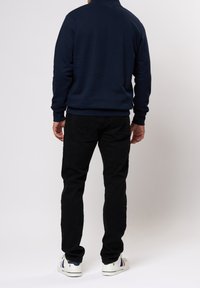 Mörkblå tröja med hög krage, kombinerad med svarta jeans och vita sneakers med en färgad rand på sidan.