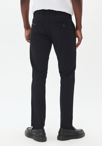 Zwarte pantalon met een slim fit. Voorzien van een vlakke voorkant, twee achterzakken met knopen en een gladde textuur. gedragen met zwarte schoenen.