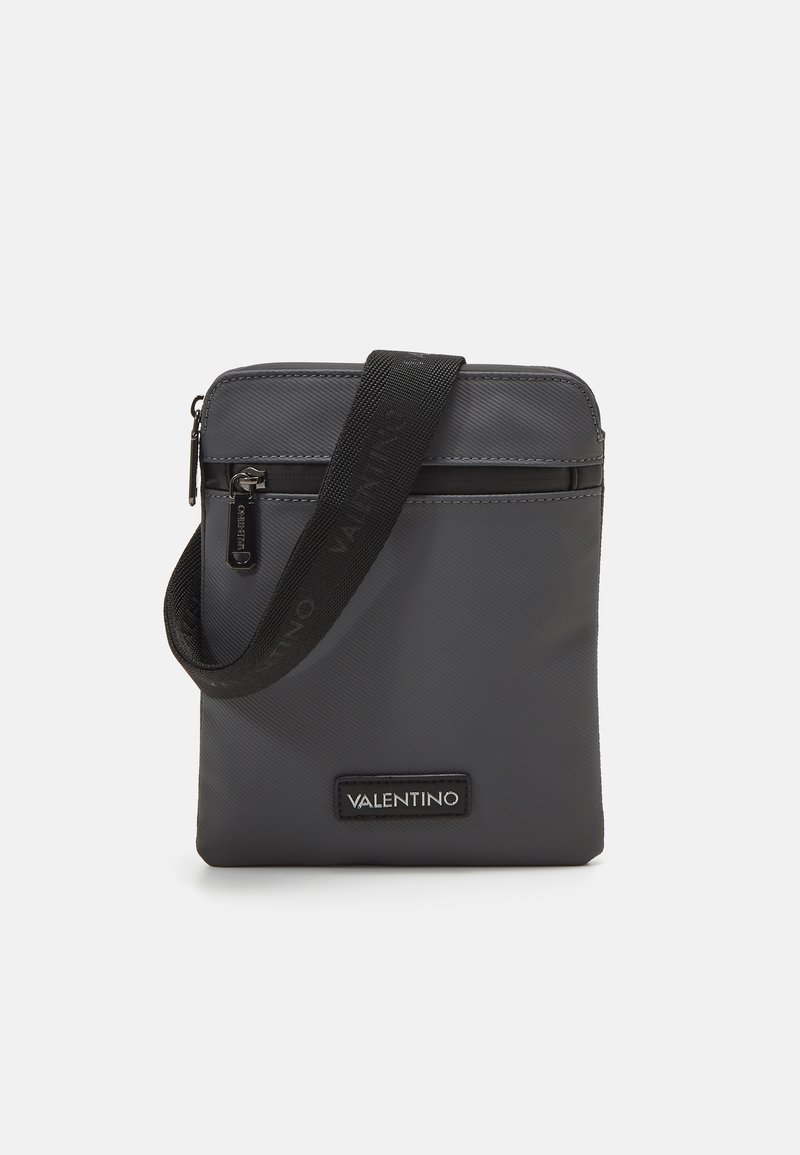 Grå nylon crossbody-väska med dragkedja, justerbar svart rem och en märkeslogopatch på framsidan. Slät textur, minimalistisk design.