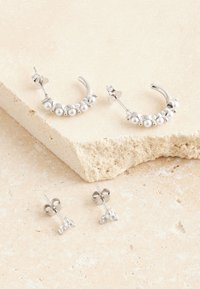 Silberne Creolen mit kleinen weißen Perlen und Ohrsteckern mit Kristallakzenten, auf einer strukturierten, hellen Steinoberfläche platziert.