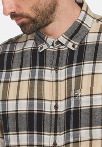 Geruite button-up shirt in beige, zwart en wit. Heeft een spread collar, houten knopen en een klein logo op de borstzak. Zachte textuur.