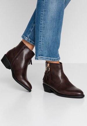 Bottines en cuir marron avec un petit talon carré, fermeture éclair sur le côté et accents de pompons décoratifs, associées à un jean bleu retroussé.