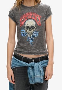 Camiseta gráfica de algodón en gris deslavado con un diseño de calavera, texto en rojo y azul, y un corte corto. Combinada con un cinturón negro y un envoltorio de mezclilla.