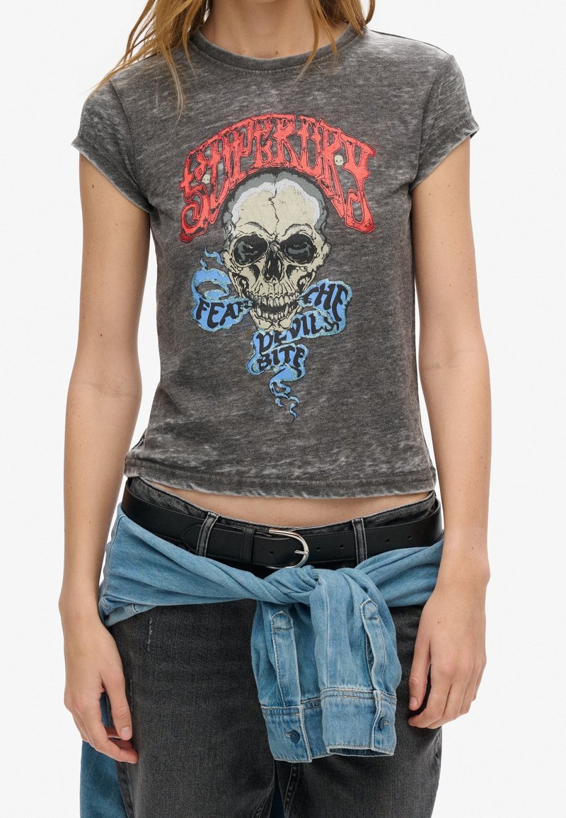 Camiseta gráfica de algodón en gris deslavado con un diseño de calavera, texto en rojo y azul, y un corte corto. Combinada con un cinturón negro y un envoltorio de mezclilla.